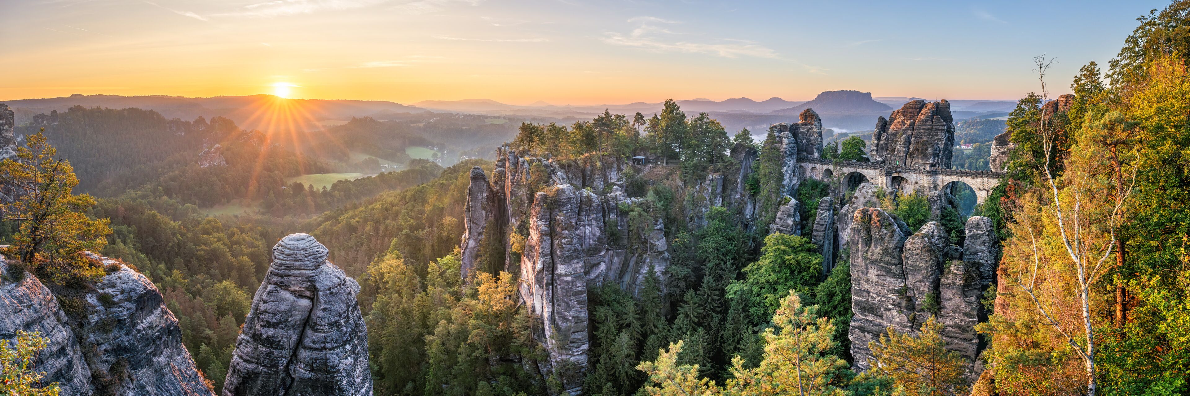 A forested landscape with prominent rock formations and a distant view of a sunset.