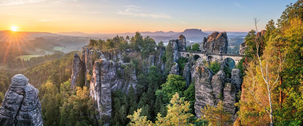 A forested landscape with prominent rock formations and a distant view of a sunset.