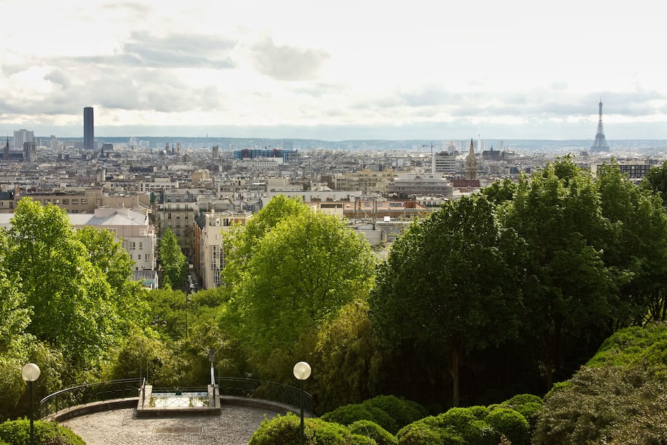 A cityscape with a prominent tower in the distance, surrounded by lush green trees and a cobblestone plaza.