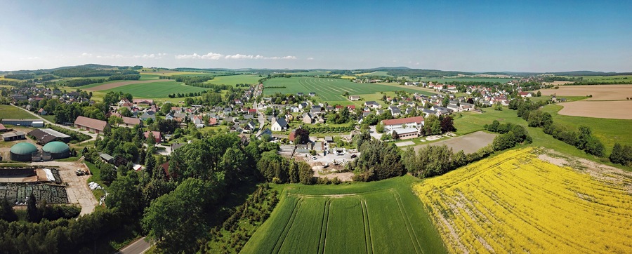Eine Vogelperspektive auf ein ländliches Dorf mit grünen Feldern, einem dichten Häuserkern und einer großen, grünen Kuppelstruktur.