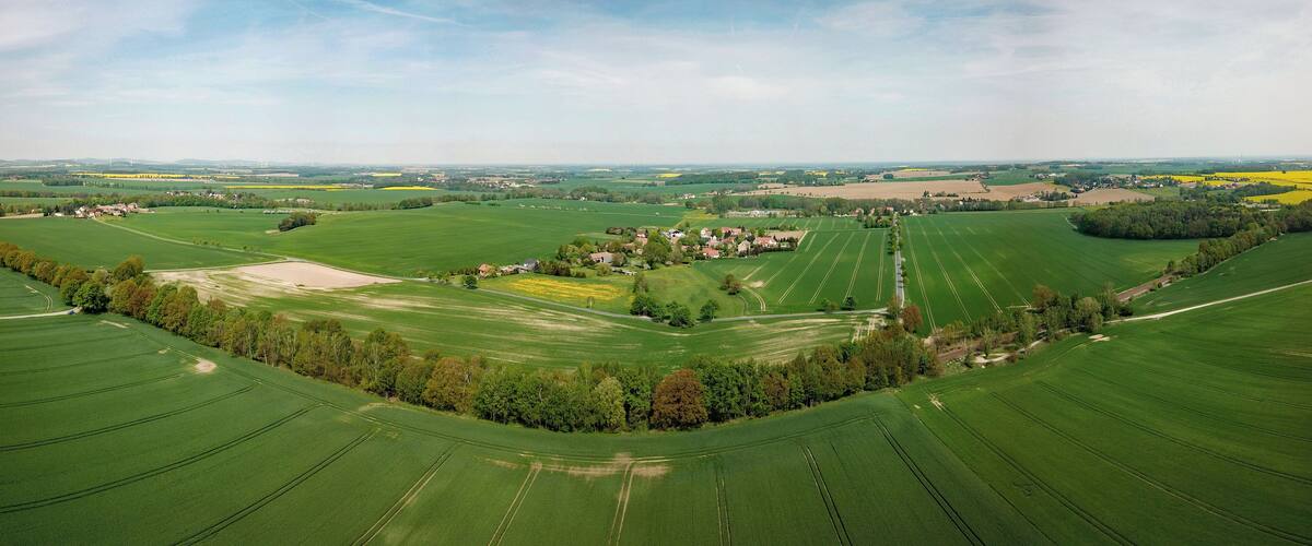 A vast agricultural landscape with neatly arranged fields, a small cluster of houses, and distant wind turbines.