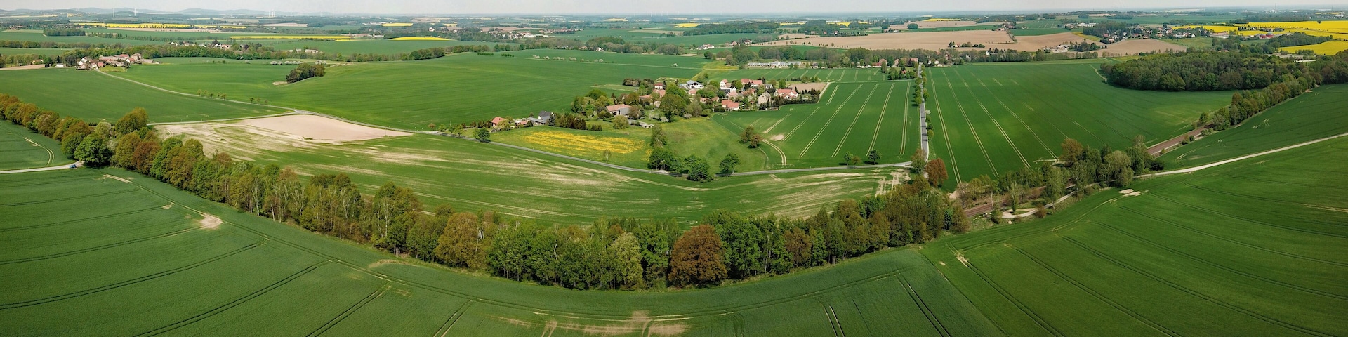 A vast agricultural landscape with neatly arranged fields, a small cluster of houses, and distant wind turbines.