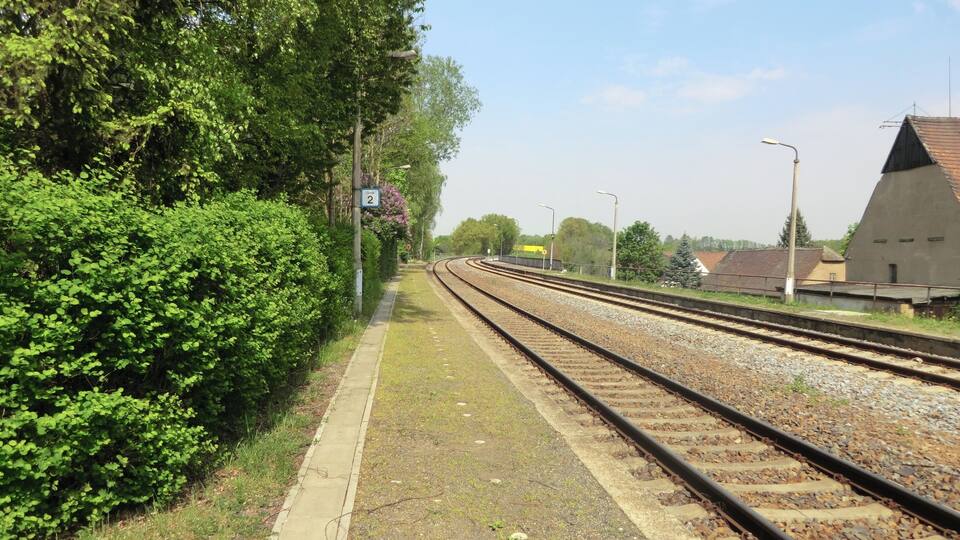A railway track with a platform, surrounded by greenery and a building with a sloped roof.