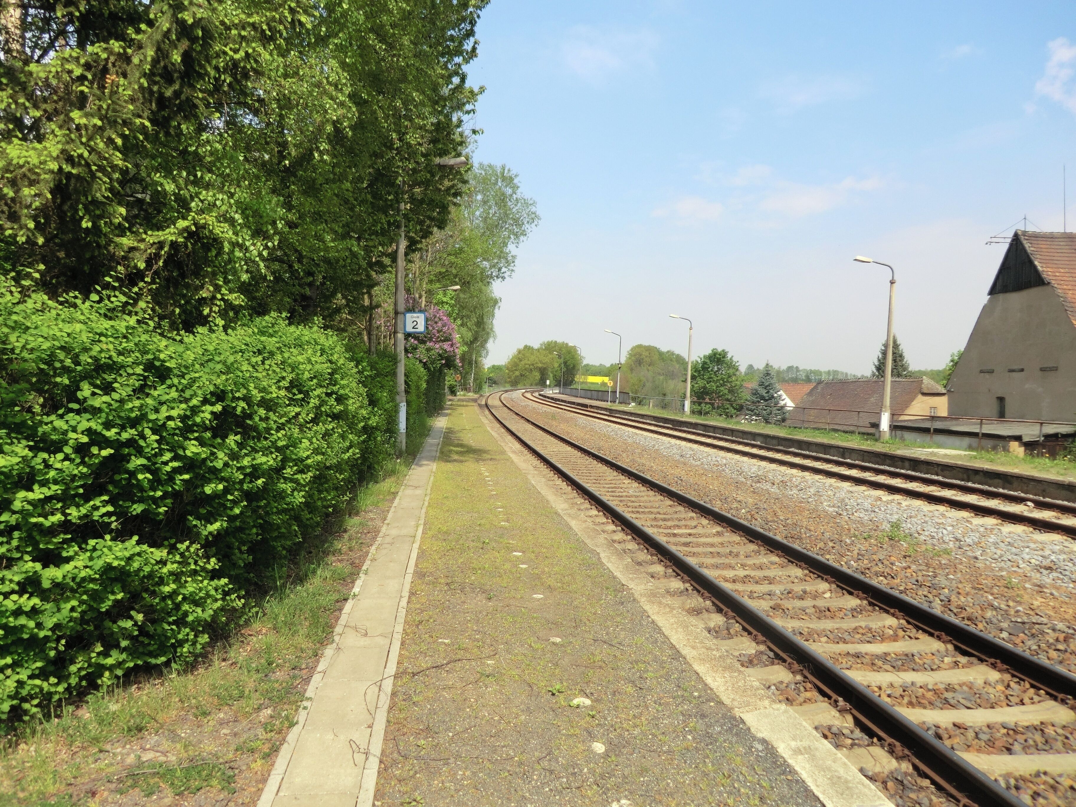 A railway track with a platform, surrounded by greenery and a building with a sloped roof.