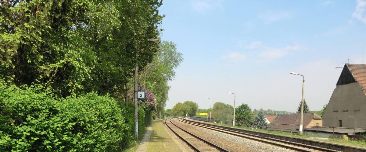 A railway track with a platform, surrounded by greenery and a building with a sloped roof.