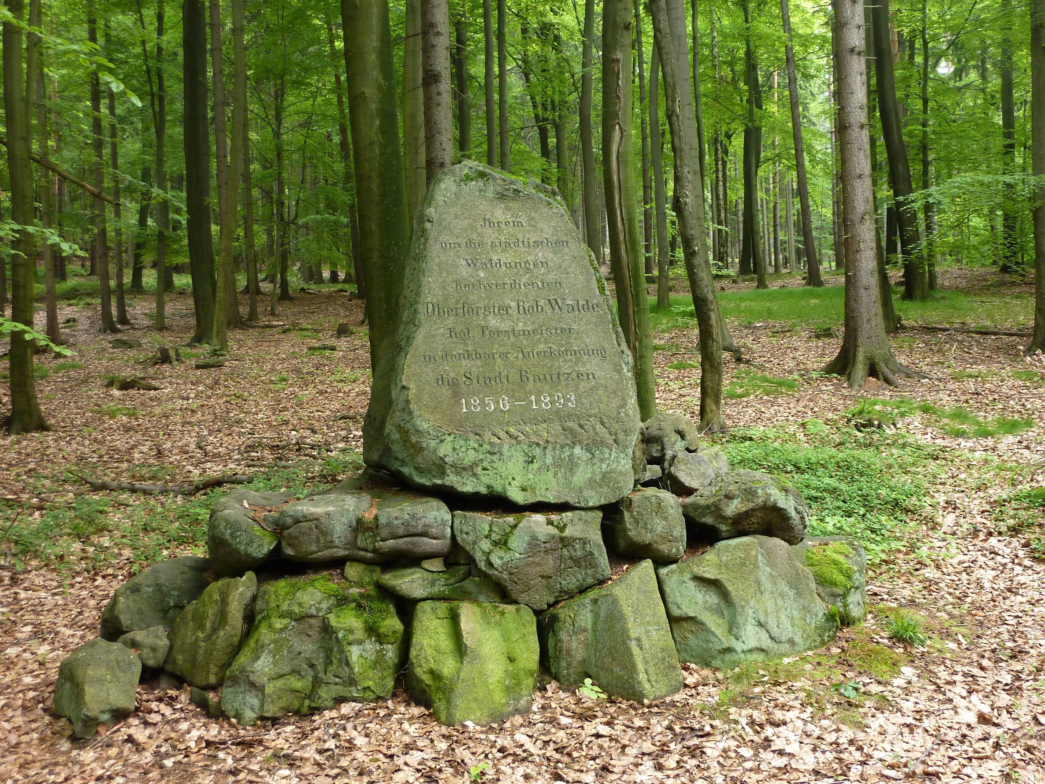 A stone monument in a forest with inscriptions and a pile of stones at its base.