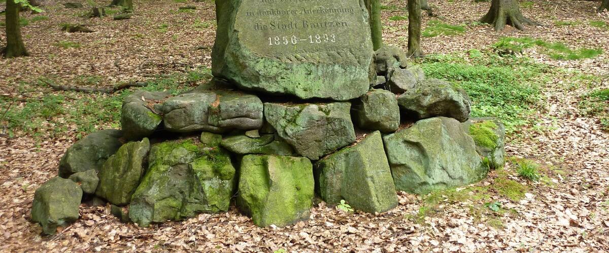 A stone monument in a forest with inscriptions and a pile of stones at its base.