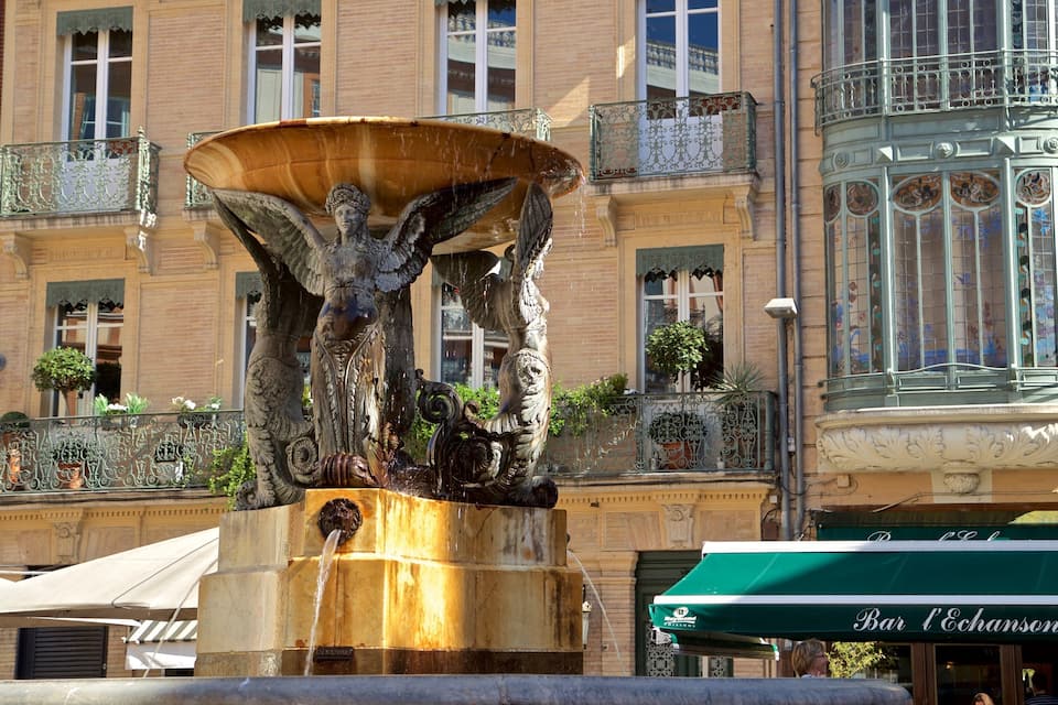 A fountain with a statue of a woman and a lion, surrounded by ornate balconies and a building with a green awning labeled "Bar l'Echanson".