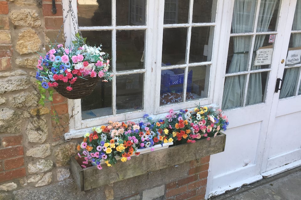 Un edificio con una puerta blanca, una ventana con una cesta de flores, y una jardinera llena de flores coloridas.