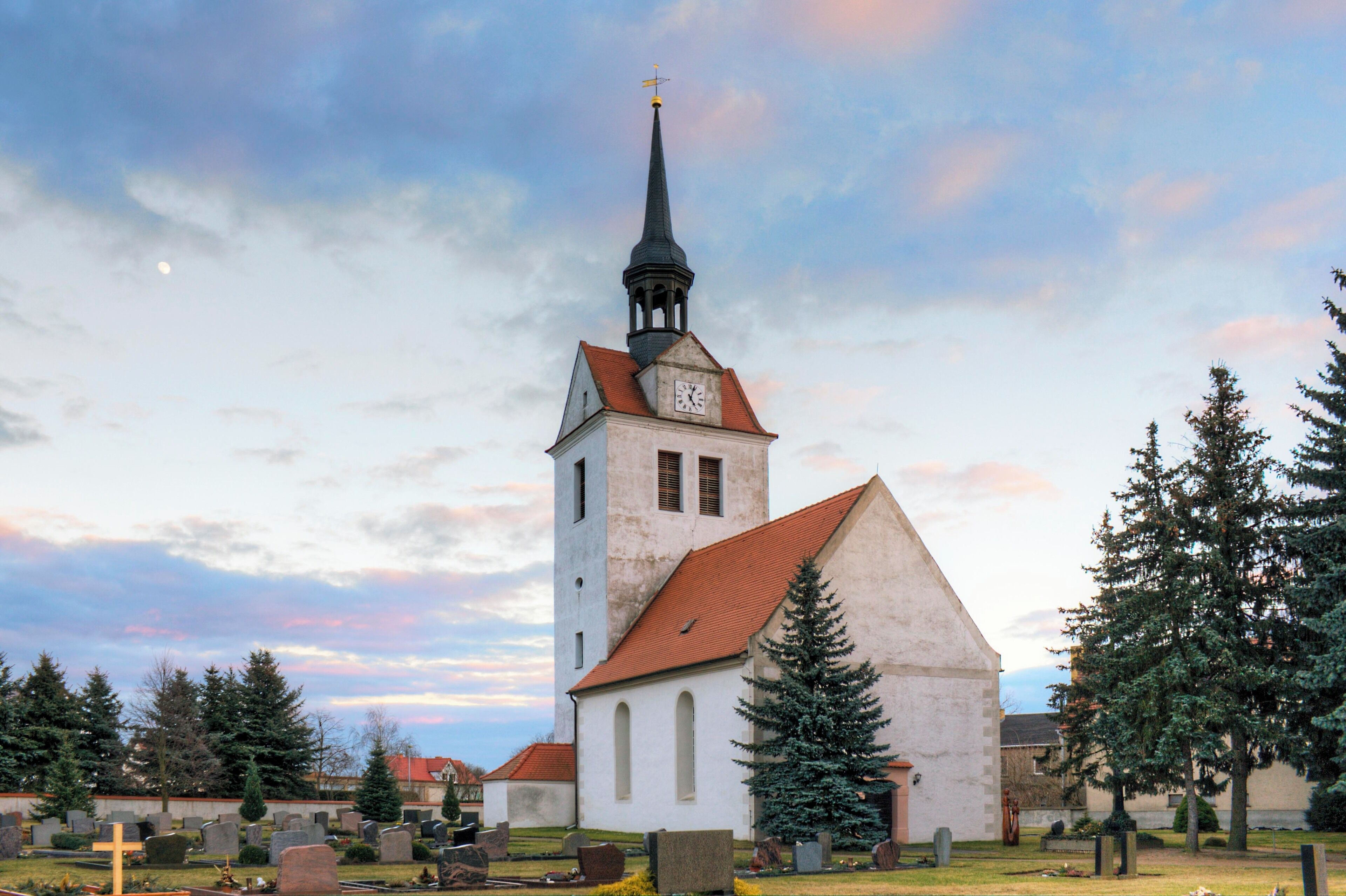 A church with a tall steeple and a clock tower, surrounded by gravestones and trees.