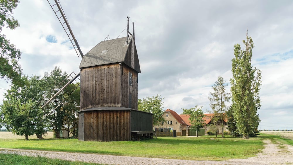 Un moulin à vent en bois, doté d’un treuil métallique, entouré d’arbres et d’une maison au toit rouge.