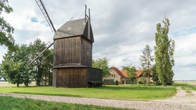 A wooden windmill with a metal windlass, surrounded by trees and a house with a red roof.