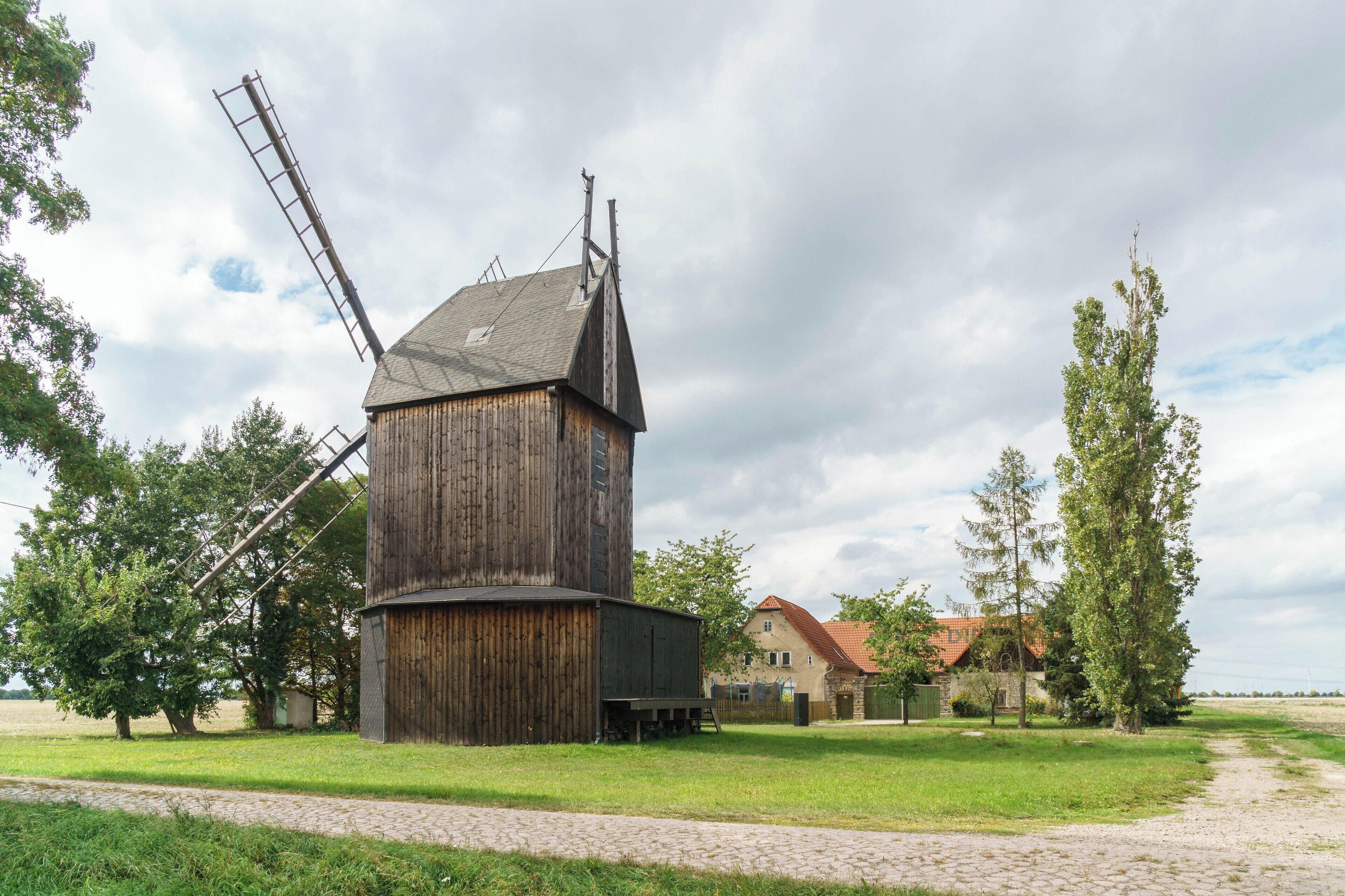 A wooden windmill with a metal windlass, surrounded by trees and a house with a red roof.