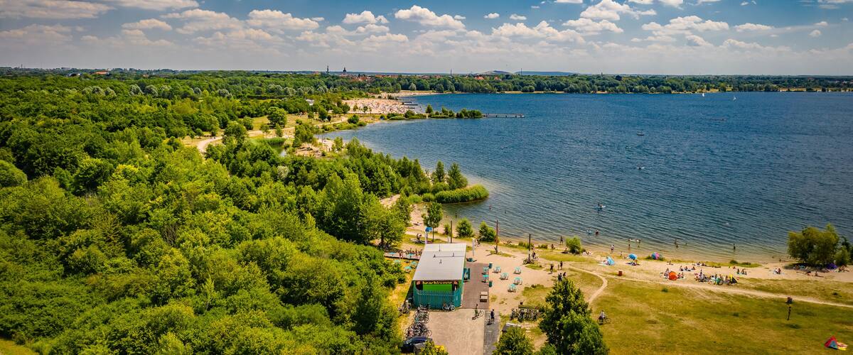 A lake surrounded by dense forest, a beach with people and tents, and a distant city skyline.