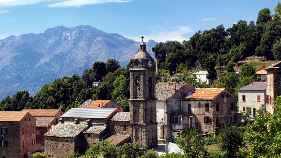 A historic church with a bell tower, surrounded by traditional stone buildings and lush greenery, with mountains in the background.