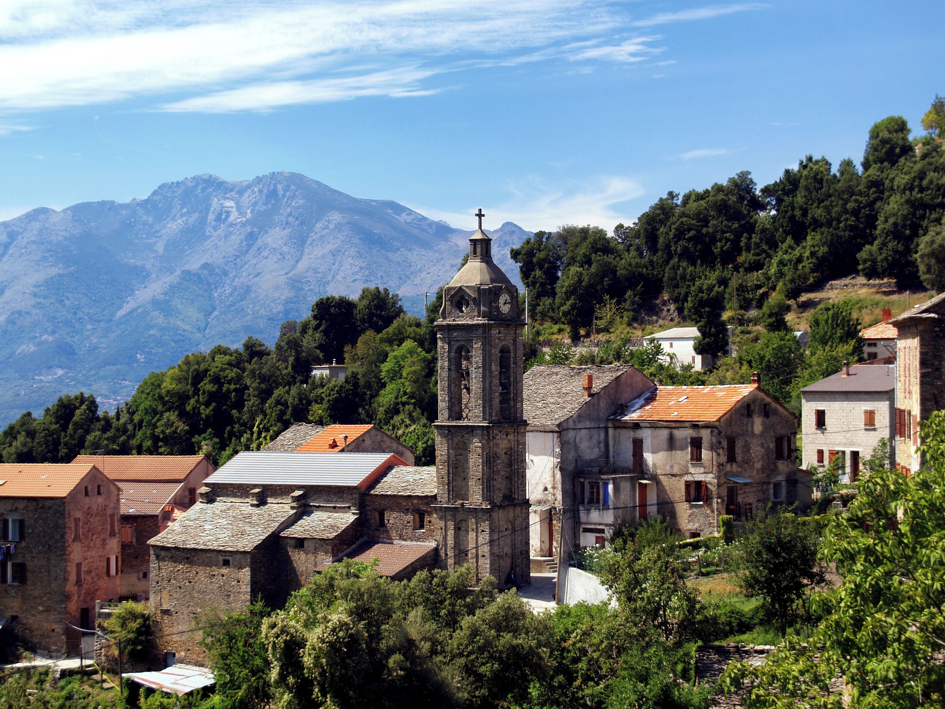 A historic church with a bell tower, surrounded by traditional stone buildings and lush greenery, with mountains in the background.