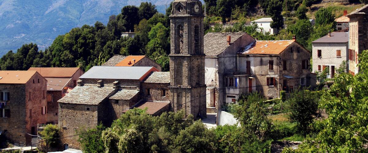 A historic church with a bell tower, surrounded by traditional stone buildings and lush greenery, with mountains in the background.