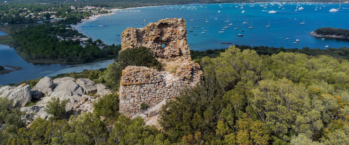 A coastal landscape with a prominent stone ruin, a bay with boats, and a distant town.