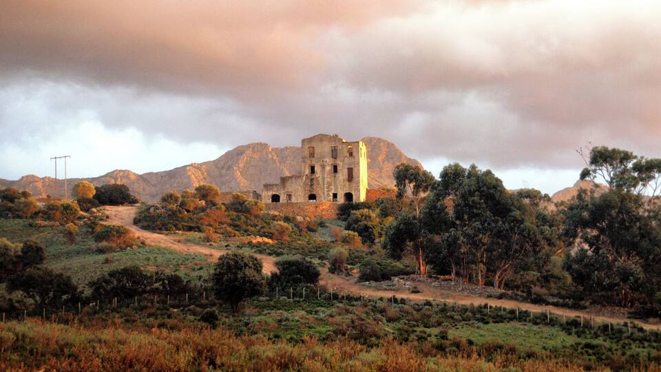 A historic stone building with arched windows, surrounded by a hilly landscape with sparse vegetation and a cloudy sky.