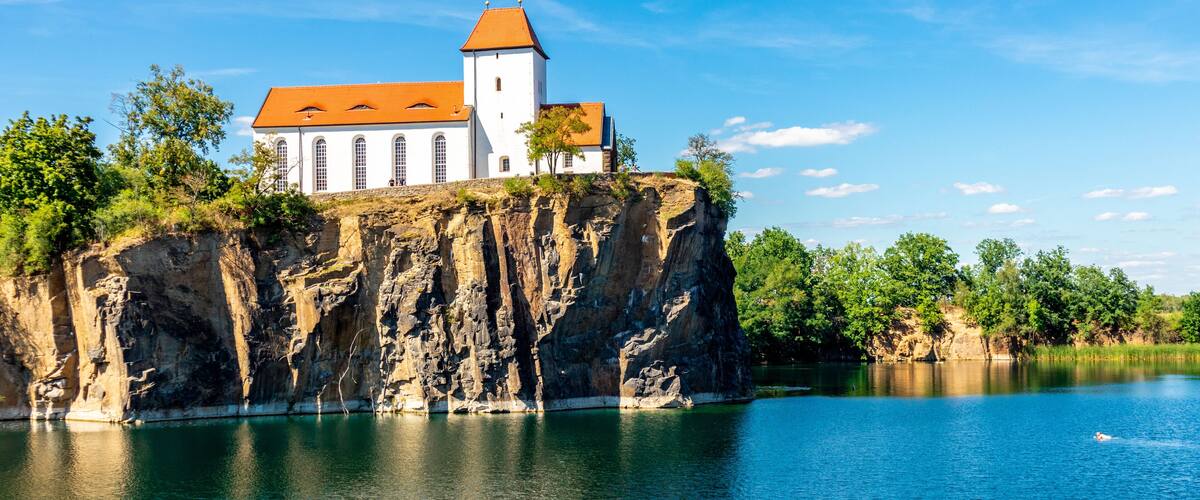 A white church with a red roof perched on a rocky cliff overlooking a calm lake.