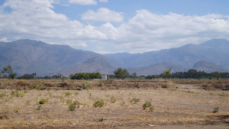 A dry, open field with scattered vegetation and a small building in the distance, surrounded by mountains.