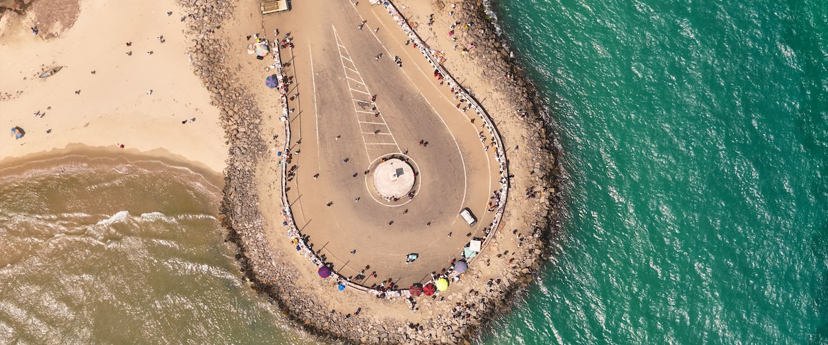 An aerial view of a beach with a circular stone structure surrounded by umbrellas and people.
