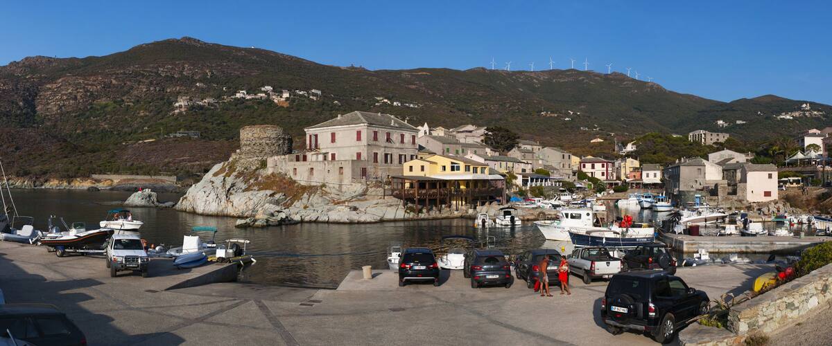 A coastal town with a marina, parked cars, boats, and historic buildings on a hillside.