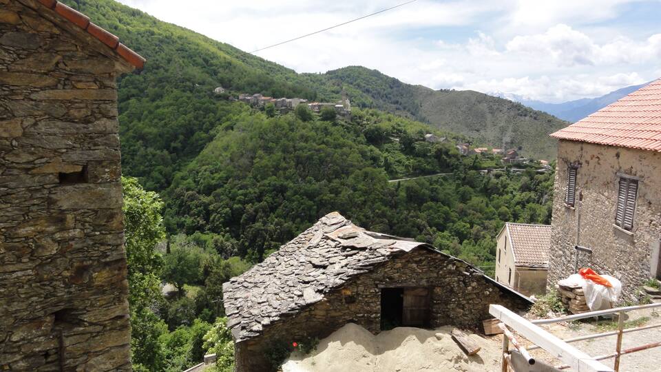 A stone building with a sloped roof, surrounded by lush green hills and a village in the distance.