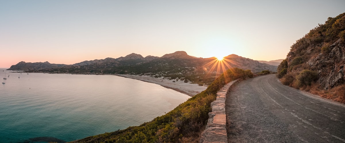 A coastal road curving along a beach with boats in the water and mountains in the background.