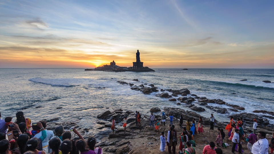 A rocky shoreline with a lighthouse and a temple in the distance during sunset.