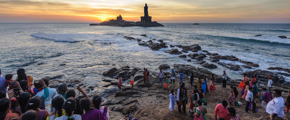 A rocky shoreline with a lighthouse and a temple in the distance during sunset.