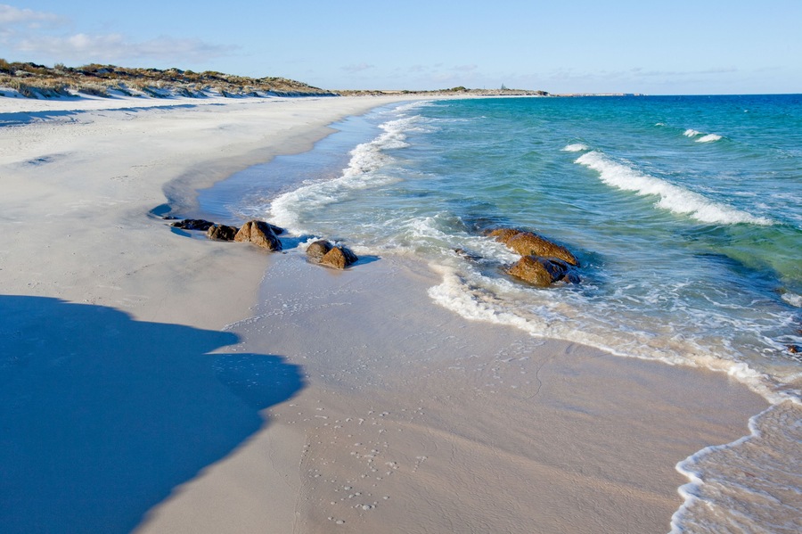 A sandy beach with clear blue water, rocks, and dunes.