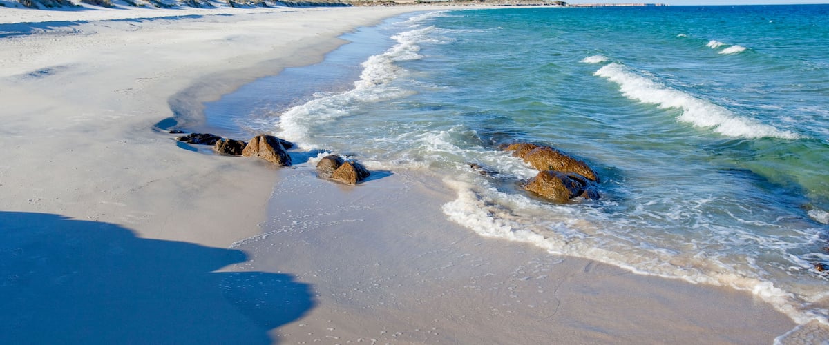 A sandy beach with clear blue water and rocks in the foreground.
