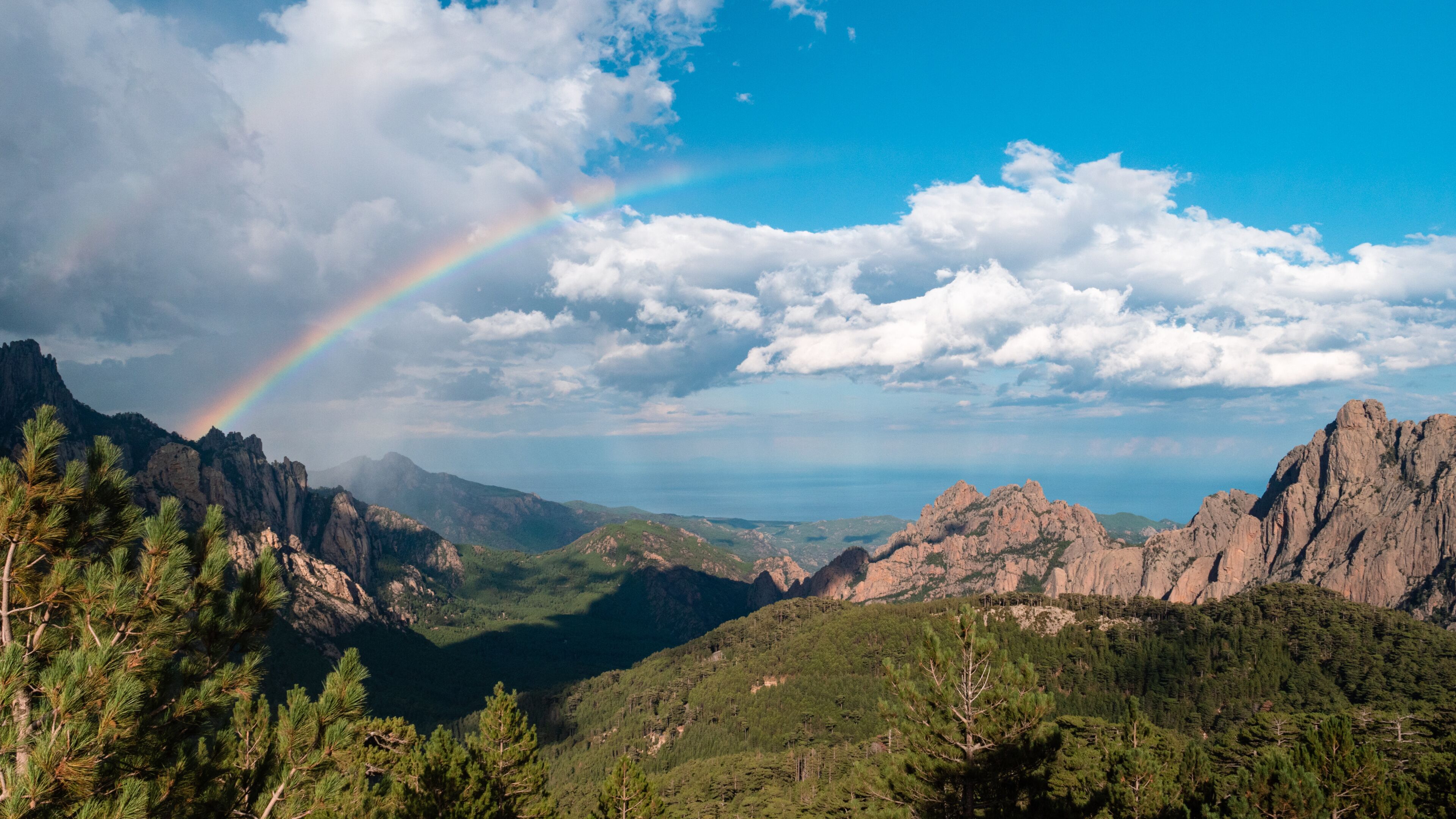 A mountain landscape with a rainbow, dense forests, and rocky outcrops.