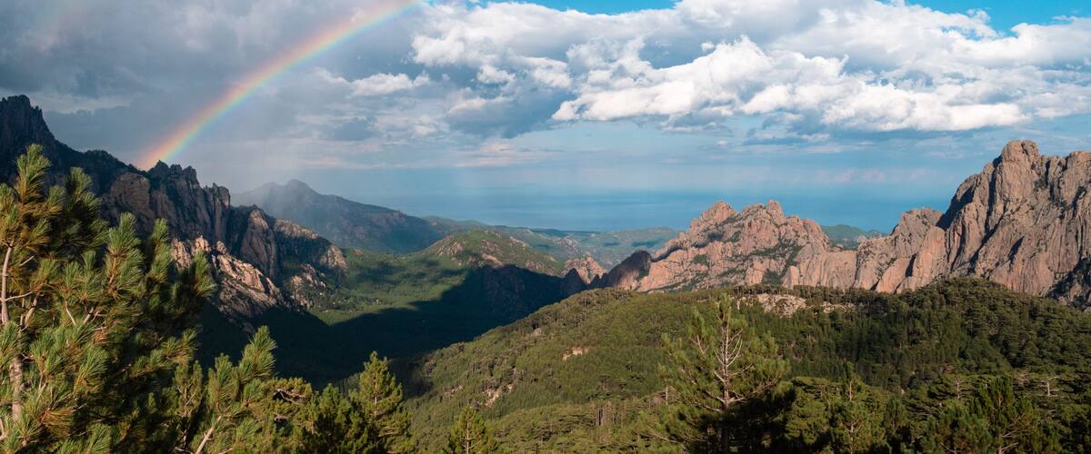 A mountain landscape with a rainbow, dense forests, and rocky outcrops.