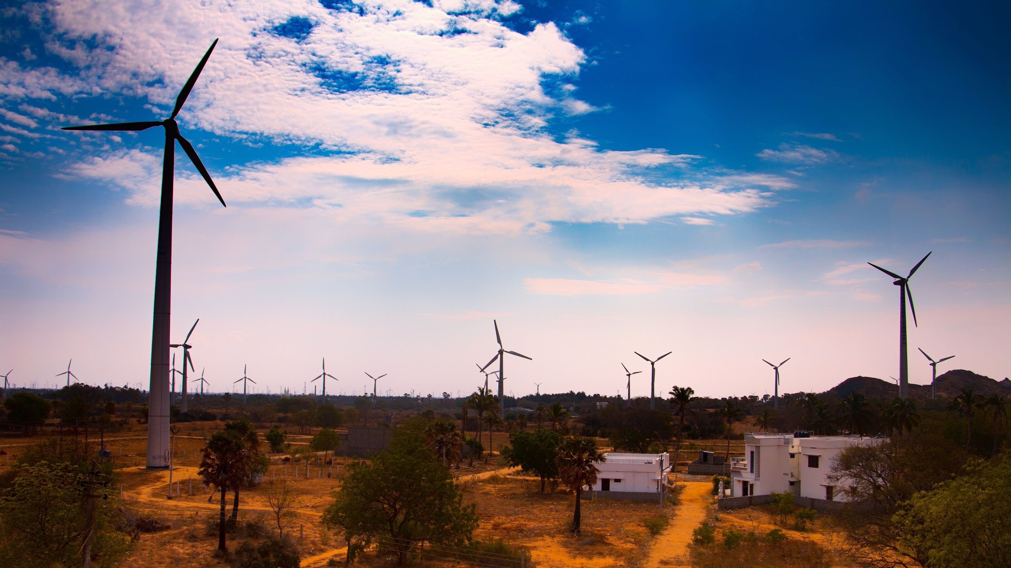 A wind farm with multiple wind turbines in a rural landscape with sparse vegetation and a few buildings.