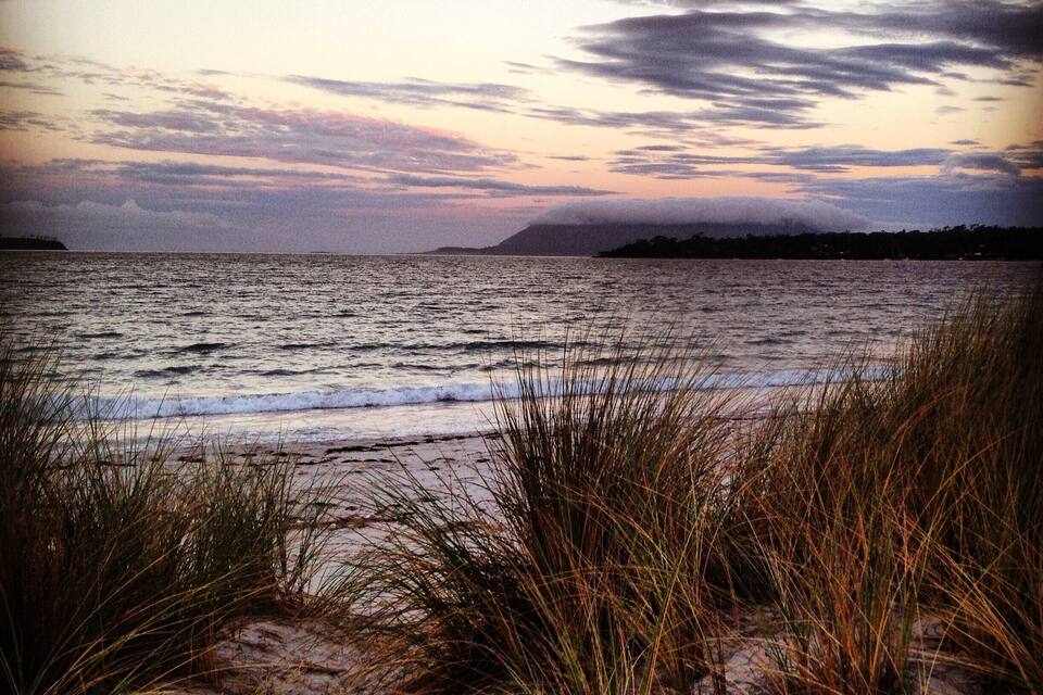 A beach with sand dunes and grass in the foreground, and a cloudy sky over the ocean.