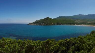 A coastal landscape with a prominent green hill, a beach, and a clear blue sea.
