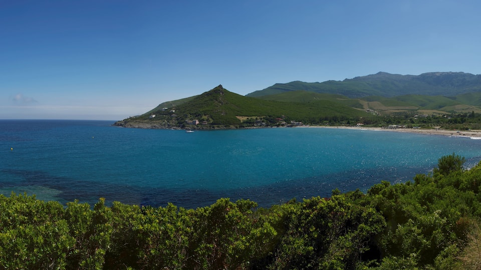 A coastal landscape with a prominent green hill, a beach, and a clear blue sea.
