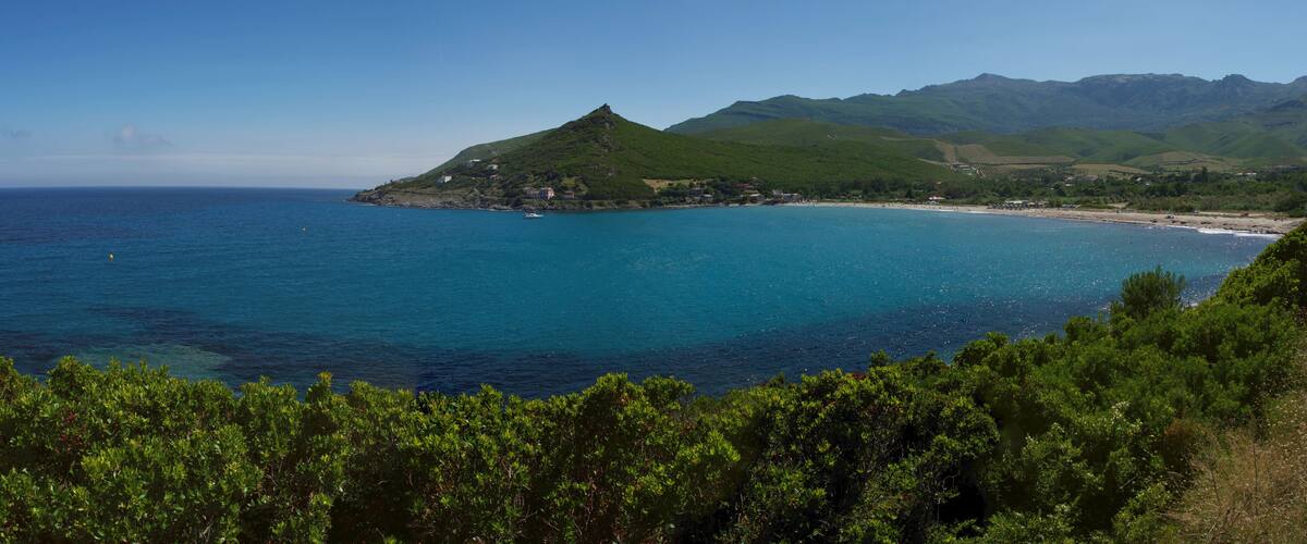A coastal landscape with a prominent green hill, a beach, and a clear blue sea.