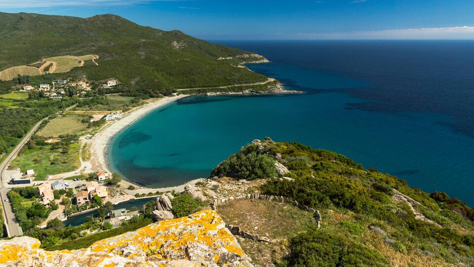 A coastal landscape with a clear blue sea, a sandy beach, and a mountainous backdrop.