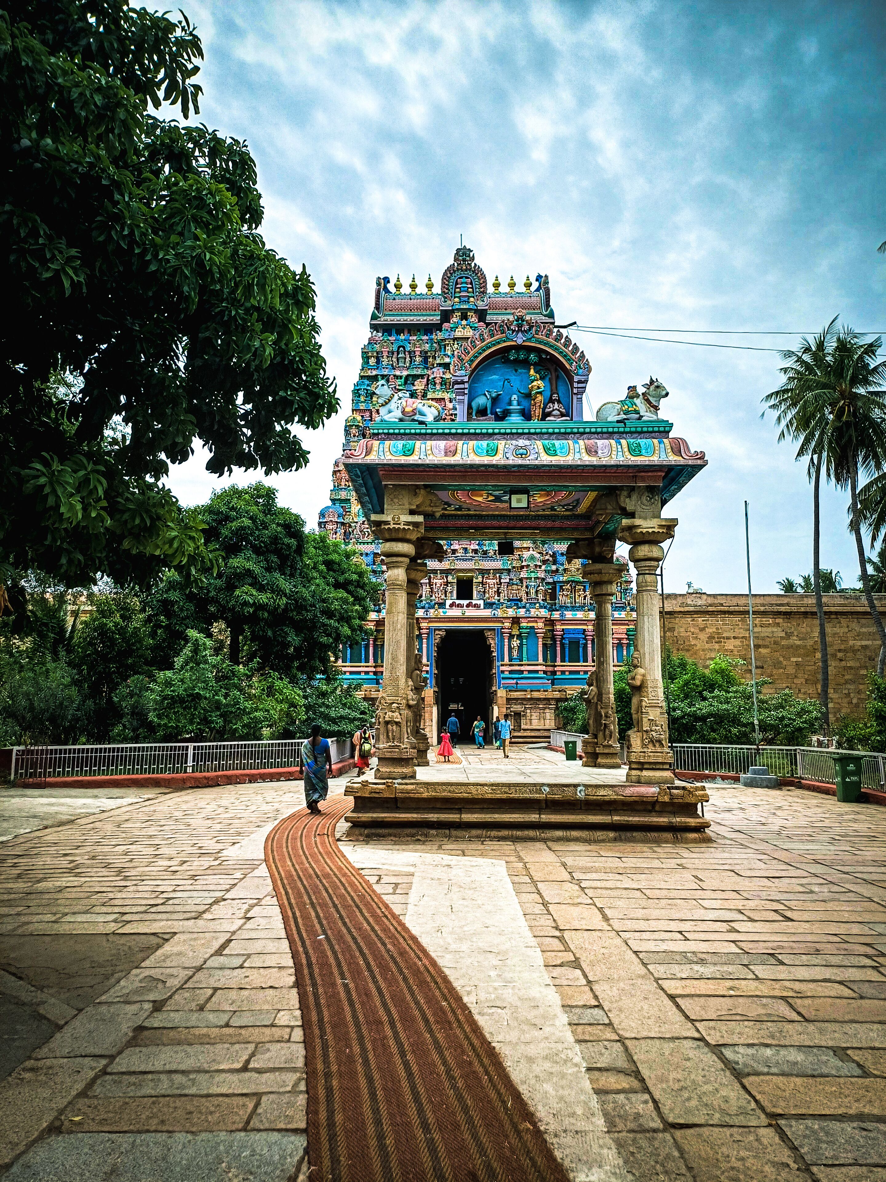 A temple with intricate carvings and statues, surrounded by trees and a paved walkway.