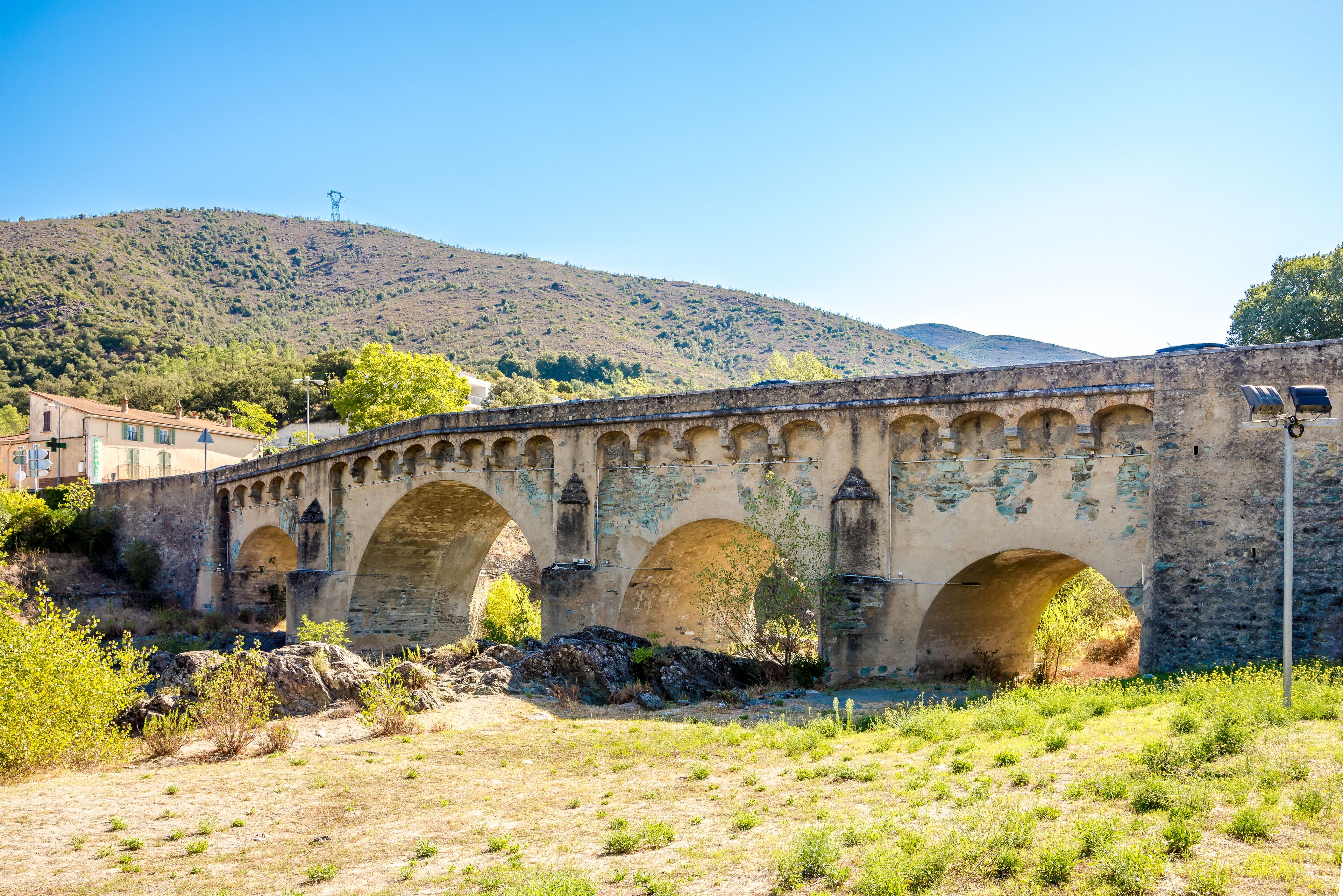 An old stone bridge with three arches over a dry riverbed, surrounded by hills and sparse vegetation.