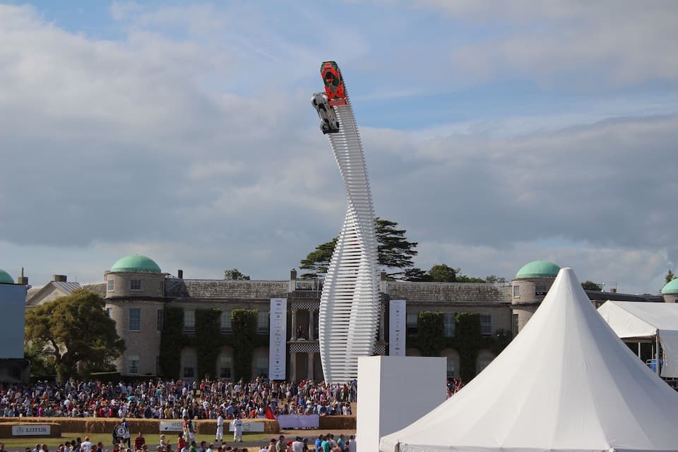 A car performing a stunt on a long white ramp in front of a crowd, with a historic building in the background.