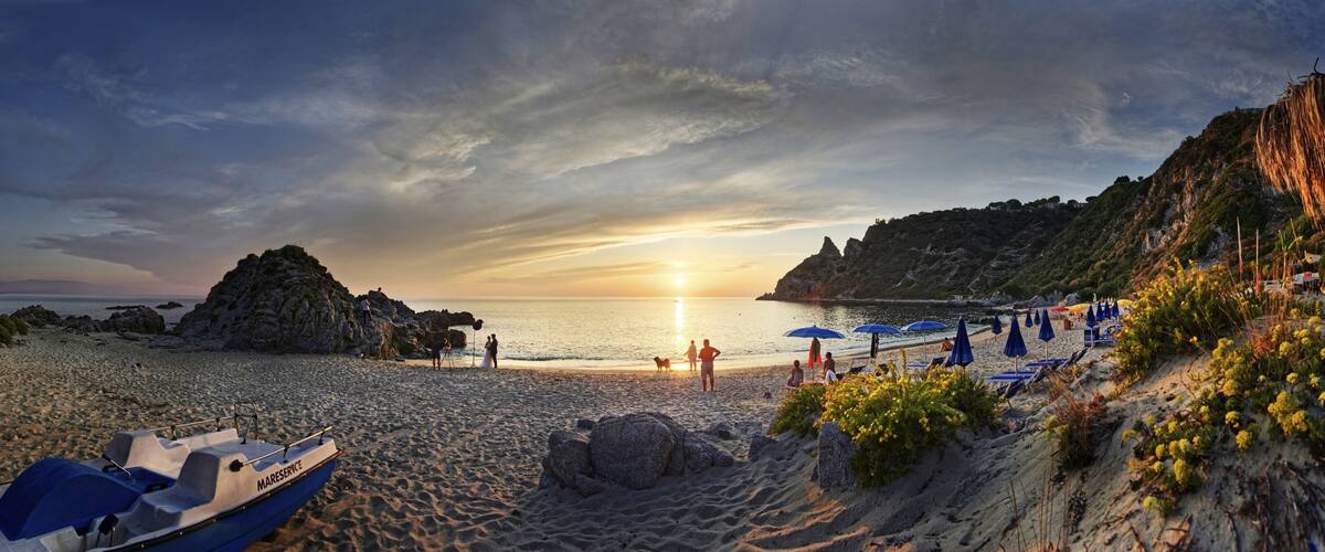 A beach with a boat, rocky cliffs, and umbrellas.