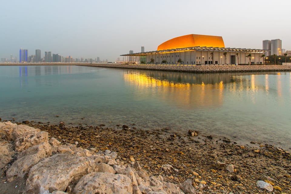 A modern building with a large orange dome, reflecting in calm water, surrounded by a rocky shoreline and cityscape in the background.