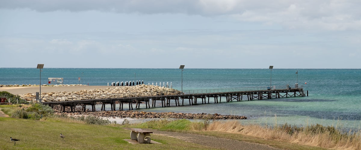 A pier extending into a calm sea, a rocky shore, and a grassy area with a bench.