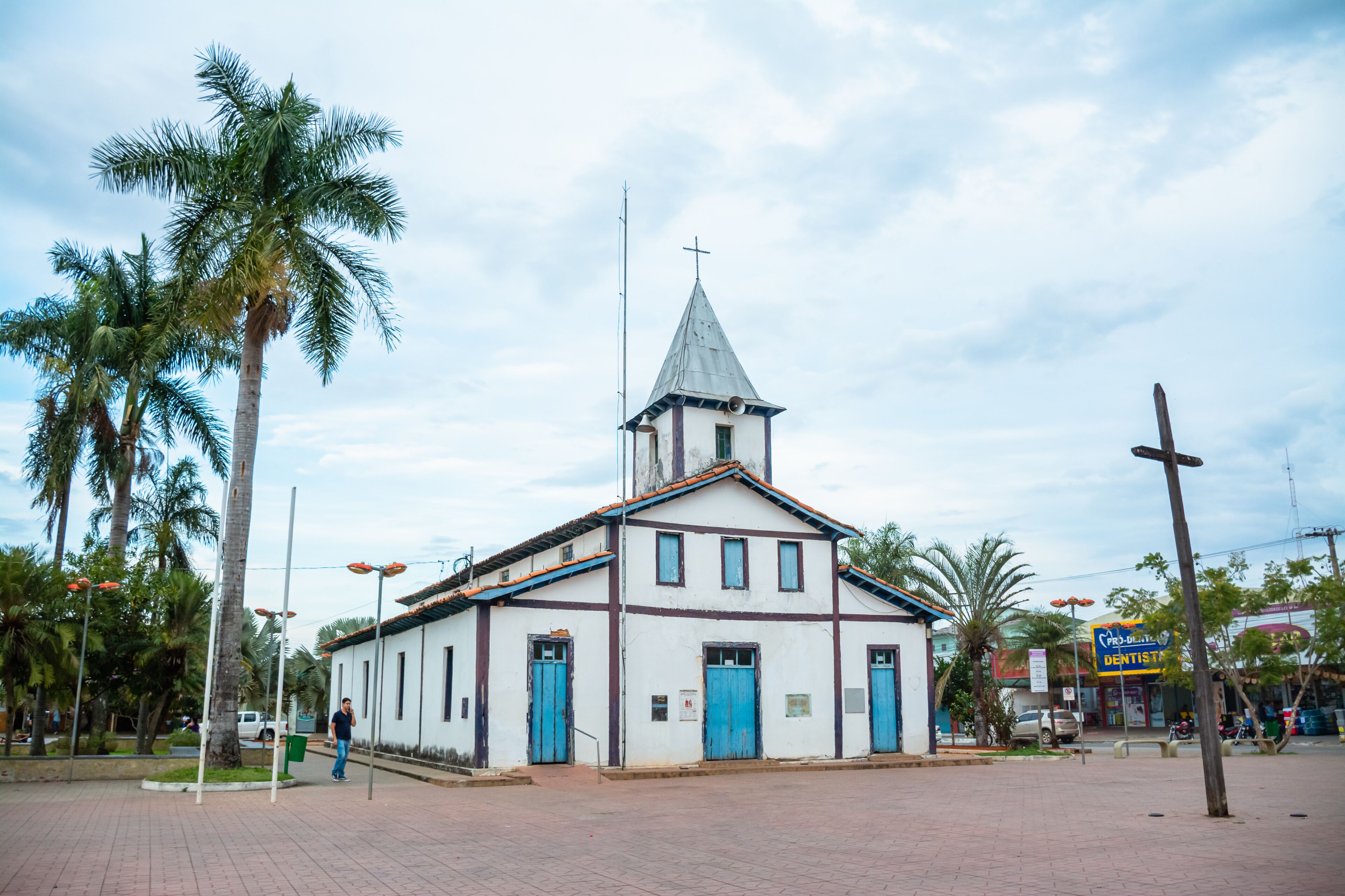 A white church with a blue door and a steeple, surrounded by palm trees and a paved area.