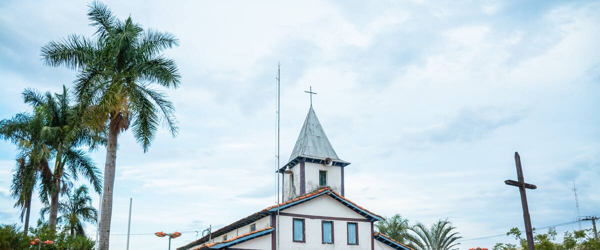 A white church with a blue door and a steeple, surrounded by palm trees and a paved area.