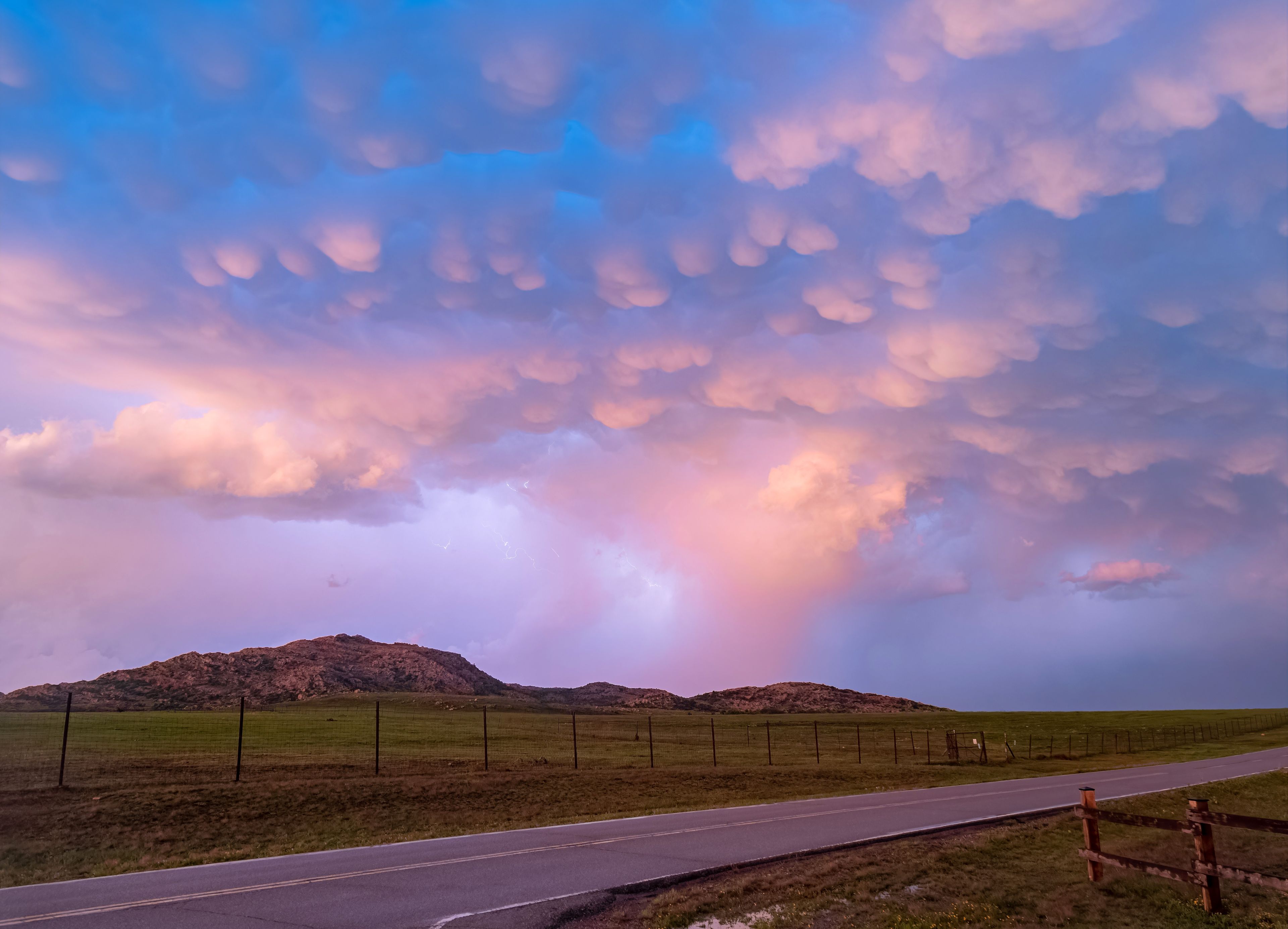 A rural landscape with a road, rolling hills, and a dramatic sky with clouds.